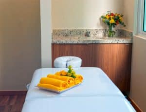 Uma sala de massagem serena do Paradise Residence com uma maca de massagem coberta por toalhas amarelas e uma bacia com flores, ao lado de uma bancada de granito com um vaso de flores frescas.