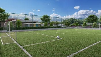 Campo de futebol society com gramado sintético, traves e uma bola no centro, cercado por uma rede e paisagem verde no DUO Residencial das Árvores.