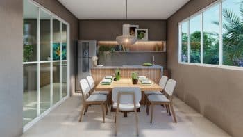 Espaço gourmet elegante no DUO Residencial das Árvores com uma mesa de jantar de madeira, cadeiras modernas e uma cozinha completa ao fundo, com uma grande janela revelando uma vista para a vegetação externa.