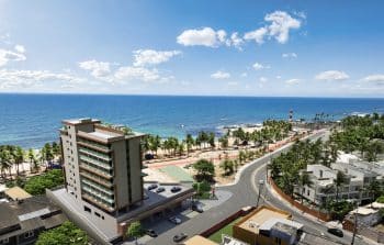 Imagem aérea mostrando um edifício moderno ao lado de uma praia tropical, com palmeiras, um farol e uma estrada curva em Salvador, Bahia.
