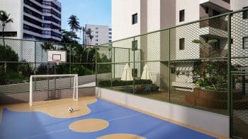 Imagem de uma quadra poliesportiva colorida e cercada por grades verdes, com um gol de futebol e uma cesta de basquete, localizada entre edifícios residenciais.