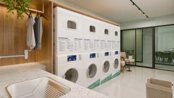 Interior de uma lavanderia equipada no BOSSA Farol de Itapoan, com várias máquinas de lavar e secar empilhadas, pia de inox e uma área de espera confortável.
