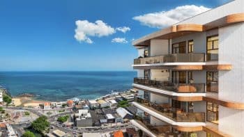 Vista lateral de um prédio moderno de vários andares com varandas de vidro e detalhes em madeira, localizado à beira-mar em uma área urbana.