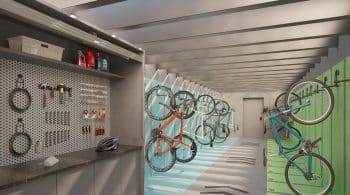 Imagem de um bicicletário interno, bem organizado, com várias bicicletas suspensas em ganchos na parede pintada de verde. Ao lado, uma bancada com ferramentas para manutenção de bicicletas, incluindo chaves, alicates e parafusos, montada em uma parede com placa perfurada para organização.