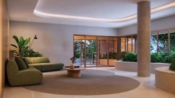 Interior do lobby do Rivê, com sofá verde-oliva em forma de L, mesa de centro circular de madeira, iluminação embutida suave no teto e amplas janelas que oferecem vista para uma área verde externa.