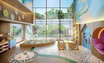 Área interna de uma brinquedoteca moderna e colorida, com escorregador transparente em espiral, piscina de bolinhas e mesas para atividades. Ao fundo, uma quadra esportiva externa.