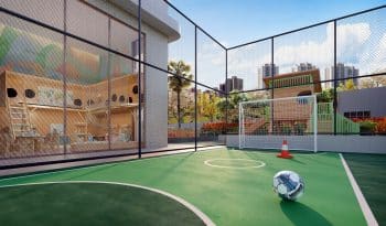 Vista de uma miniquadra esportiva com piso de concreto pintado de verde, cercada por grades de proteção, com uma bola de futebol no chão e um gol ao fundo. Ao lado, há uma área de recreação com brinquedos e vegetação.