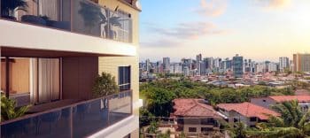 Imagem de uma varanda de um apartamento no Lodge Caminho das Árvores, com vista ampla para a cidade. A varanda possui guarda-corpo de vidro e é decorada com plantas e móveis confortáveis. Ao fundo, é possível ver a skyline da cidade com vários edifícios e uma área verde.