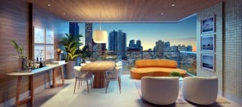 Varanda de um apartamento com decoração moderna e vista noturna da cidade. O espaço conta com uma mesa de jantar redonda, sofás confortáveis, plantas decorativas e uma parede decorada com quadros. A cidade e a Praça dos Eucaliptos são visíveis ao fundo.