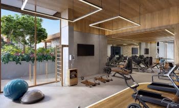 Imagem de um espaço fitness moderno, com equipamentos de musculação, esteiras, bolas de exercício e um espaldar. O ambiente tem grandes janelas que permitem a entrada de luz natural, com vista para uma área verde.