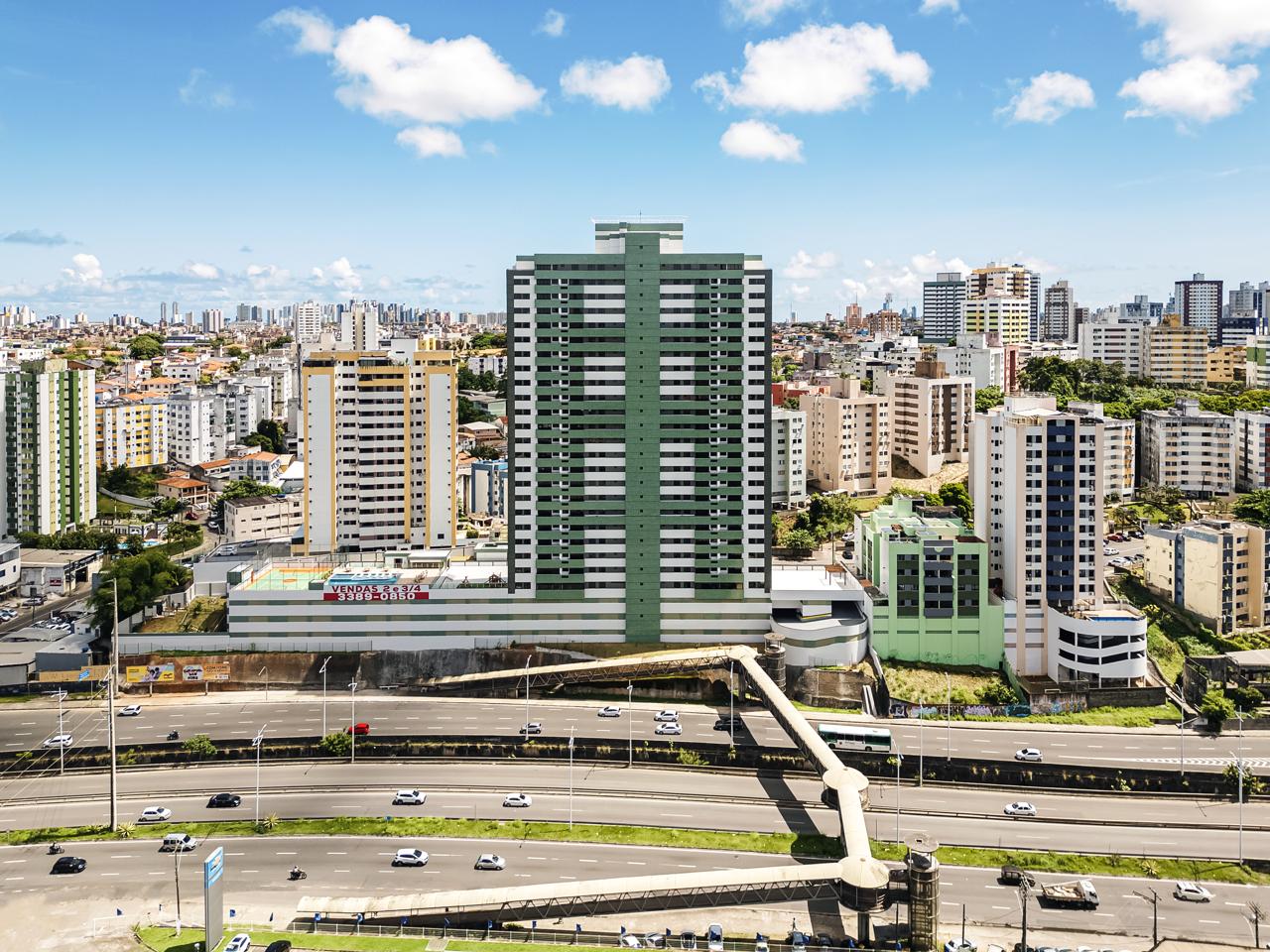 Vista aérea de um edifício residencial alto, cercado por outros prédios em uma área urbana de Salvador, com uma avenida movimentada em primeiro plano.