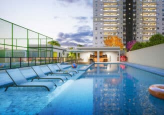 Piscina adulta com raia de 20 metros, cercada por espreguiçadeiras e área gourmet ao fundo, com quadra esportiva e torres residenciais ao redor.