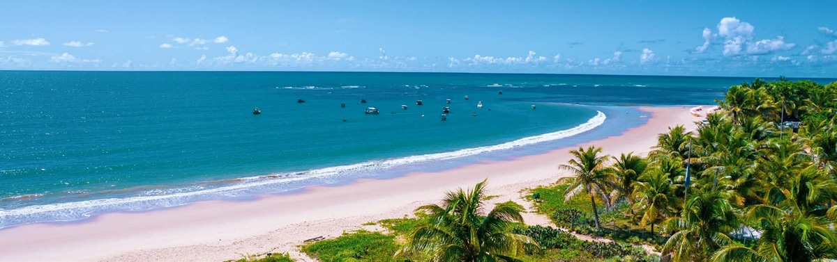 Vista panorâmica da praia de Itacimirim, com mar azul, areia clara, barcos ao fundo e uma vegetação de coqueiros na orla.