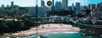 Imagem aérea mostrando a praia do Rio Vermelho em Salvador, com destaque para os edifícios modernos ao fundo e a localização do empreendimento ONE Rio Vermelho marcada.