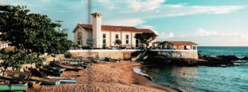 Imagem da Igreja de Santana com vista para a praia do Rio Vermelho, destacando barcos de pesca na areia e o mar tranquilo ao fundo.