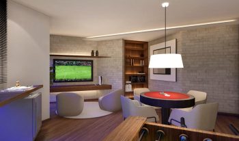 Salão de jogos do Laut Jaguaribe, com mesa de pôquer, TV de tela grande, poltronas confortáveis, estante de livros e decoração moderna em tons de madeira.