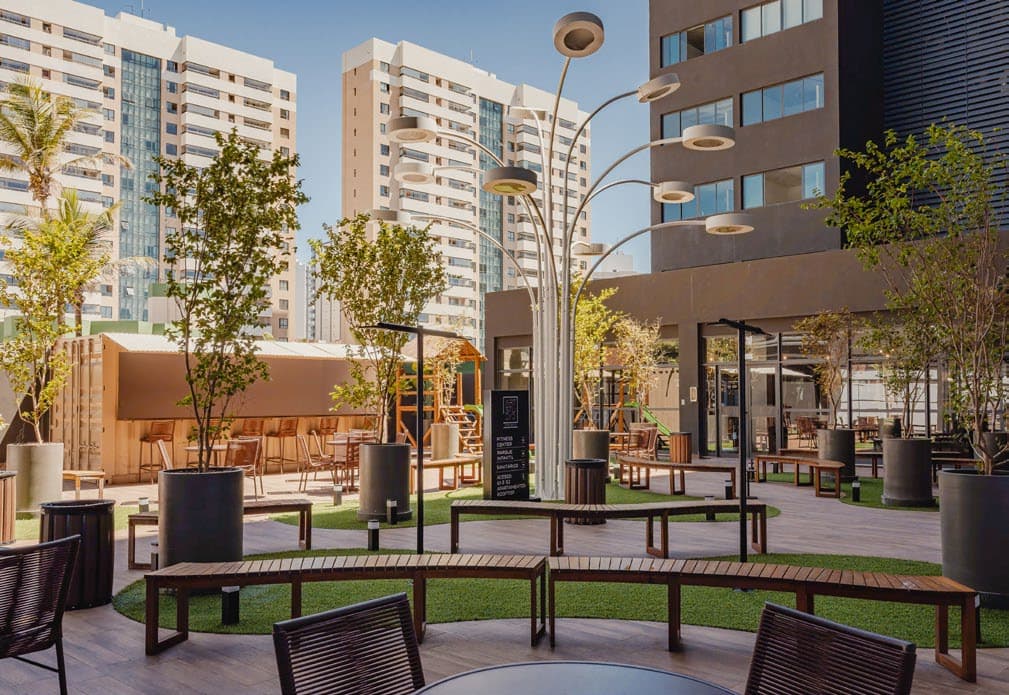 Praça de acesso do Art Studio Tancredo com bancos de madeira, árvores em vasos, iluminação moderna, áreas de convivência e prédios ao fundo.