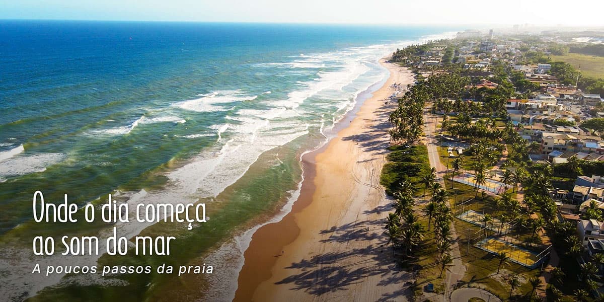 Fotografia aérea panorâmica da Praia de Stella Maris em Salvador, mostrando a faixa de areia dourada, o mar azul com ondas suaves e a orla repleta de coqueiros. À direita, vê-se a infraestrutura do bairro com ruas, casas, quadras esportivas e áreas verdes. No canto inferior esquerdo, há o texto "Onde o dia começa ao som do mar - A poucos passos da praia".